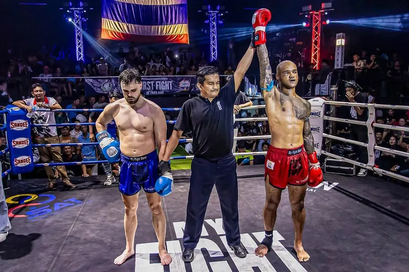 A referee raises the winner's arm after a decision, Thai flag banner and packed crowd behind.