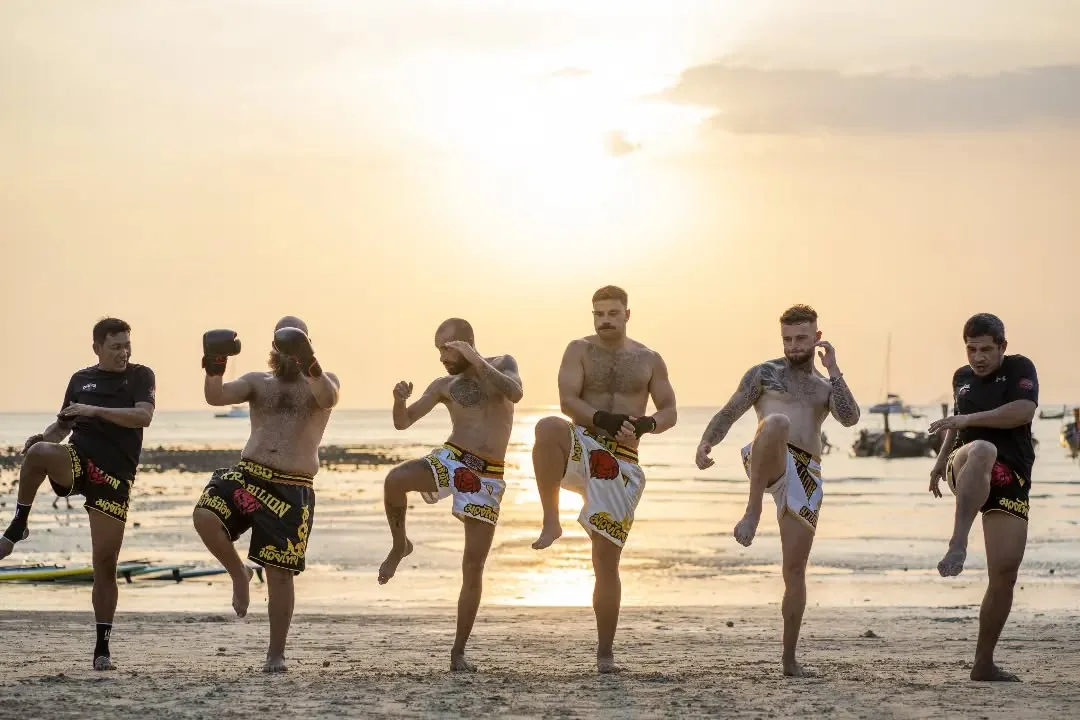 Fighters doing knee-raise drills on the beach at sunrise, karst islands and longtail boats behind.