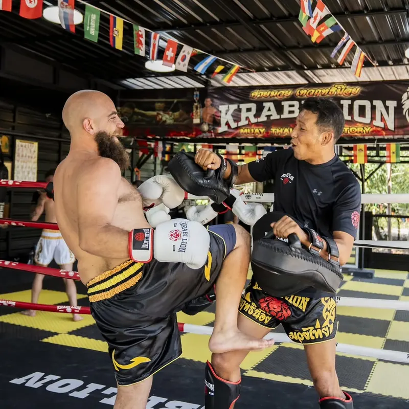 A fighter lands a full-extension roundhouse kick on the coach's Thai pads, lit from the side.
